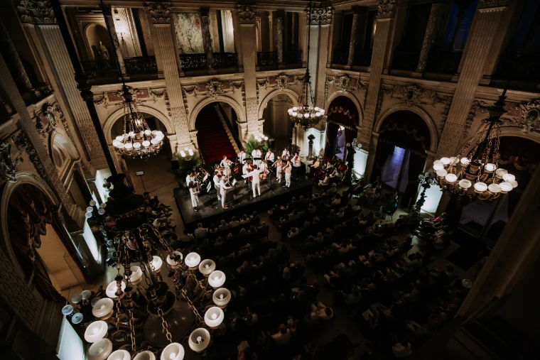 Musicians in white perform in the Great Hall of The Breakers at night, seen from the gallery (2023). Newport Classical Music Festival, The Breakers, Newport, Rhode Island, in the season calendar of The Mansa Court