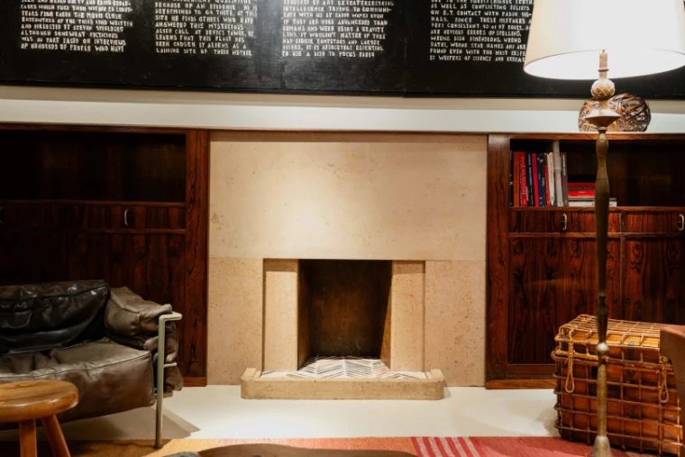 A stone fireplace set into rosewood panelling and bookshelves, with a leather armchair and a tall floor lamp, on a gallery stand (2025).