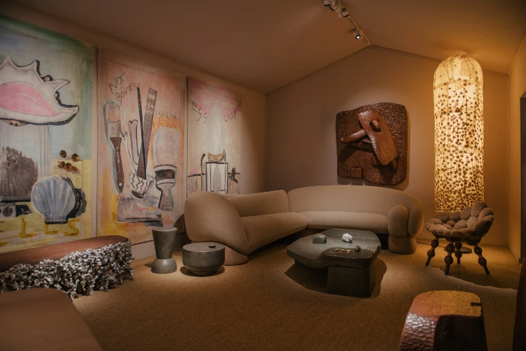 A dimly lit stand arranged as a sitting room, with a curved sofa, stone coffee table, tall glowing lamp and large painted panels on the walls (2025).