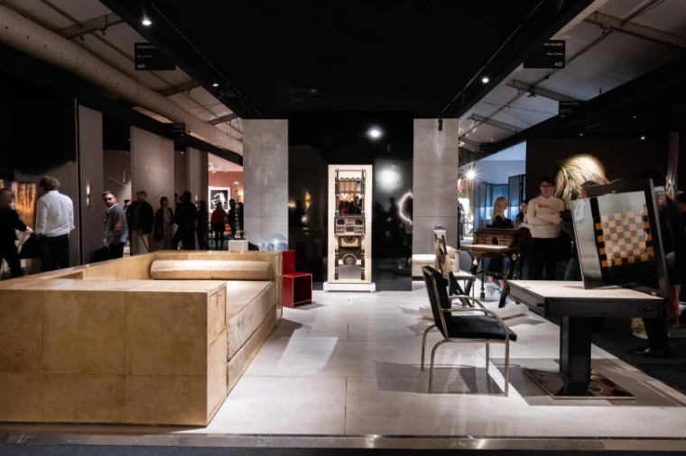 An open stand of sculptural furniture, including a low pale sofa and a chequered cabinet, with visitors in the aisles beyond (2025).