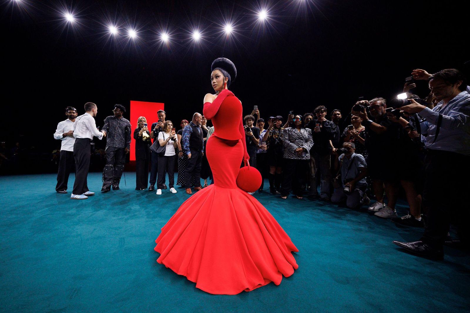 A guest in a scarlet fishtail gown turning for a bank of photographers on the teal carpet at Paris Fashion Week