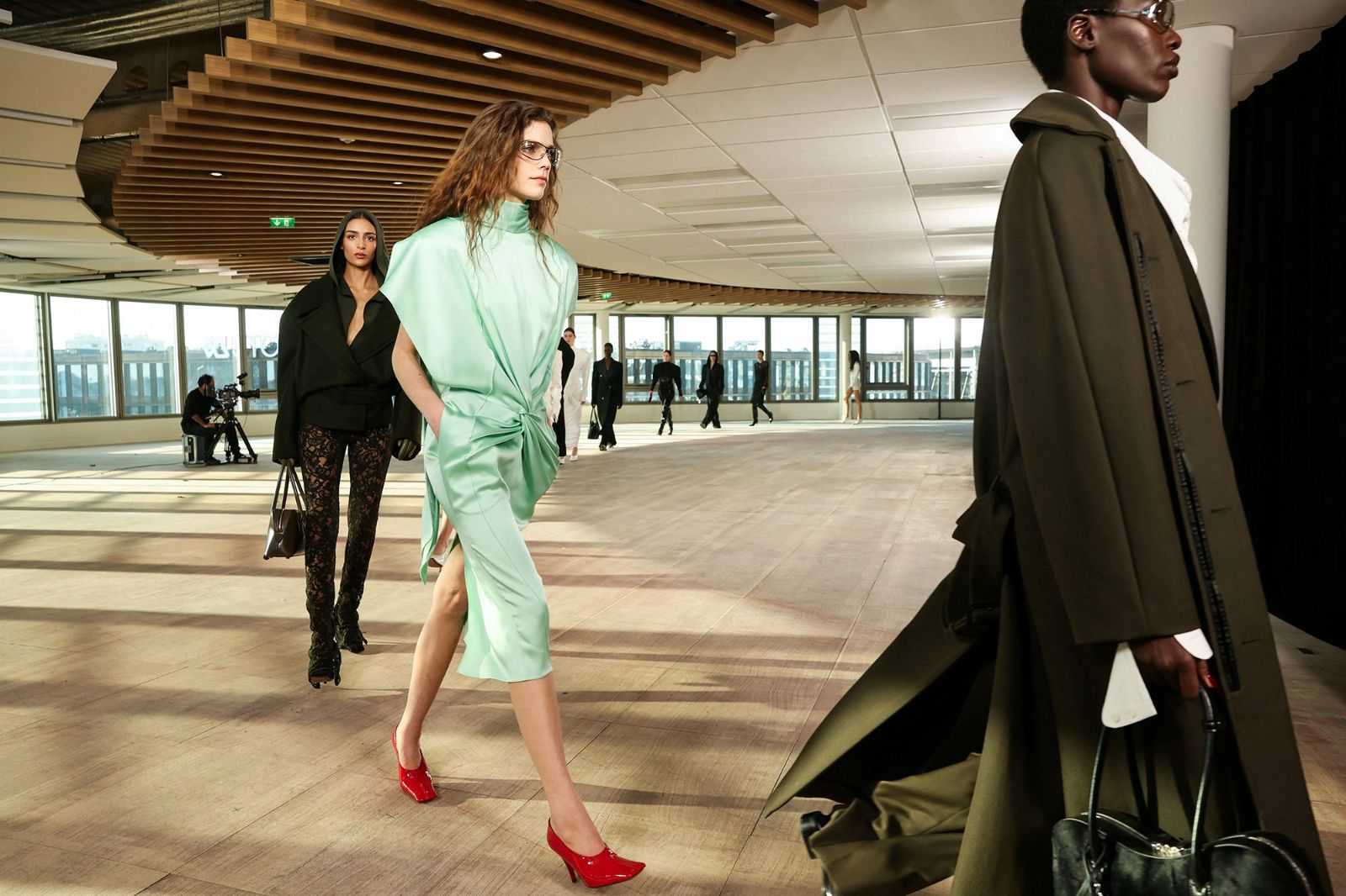 Models in tailoring and silk circling a daylit upper floor with windows onto the city at Paris Fashion Week