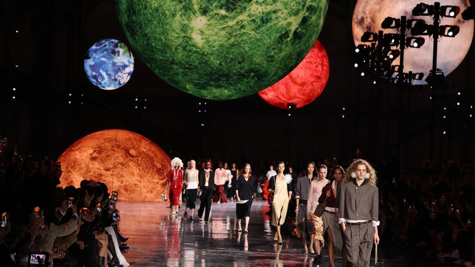 Models filing down a mirrored runway beneath vast suspended planets in a darkened hall at Paris Fashion Week