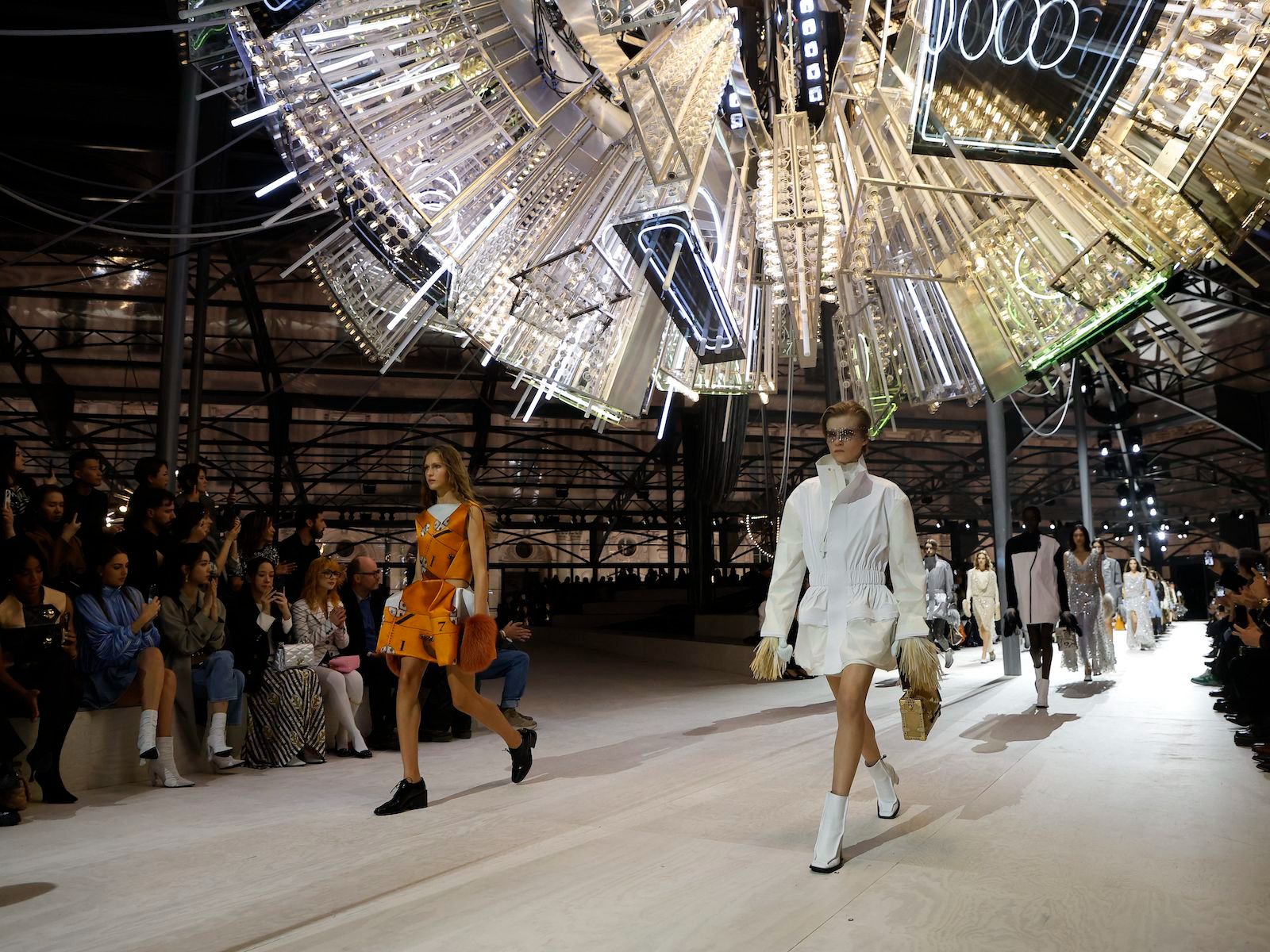 Models passing along a pale runway beneath an enormous suspended sculpture of mirrors and neon tubes at Paris Fashion Week