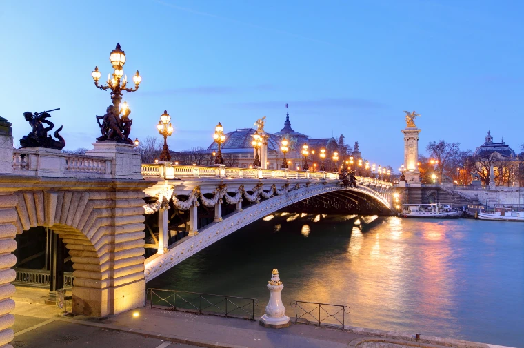 The Pont Alexandre III lit at dusk over the Seine, its lamps and gilded statues glowing, with the glass domes of the Grand Palais beyond under a blue winter sky (2015).
