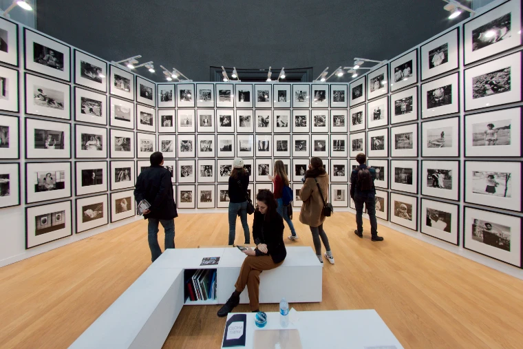 Visitors stand and sit before walls hung floor to ceiling with framed black-and-white photographs in the salon d’honneur at Paris Photo (2016).