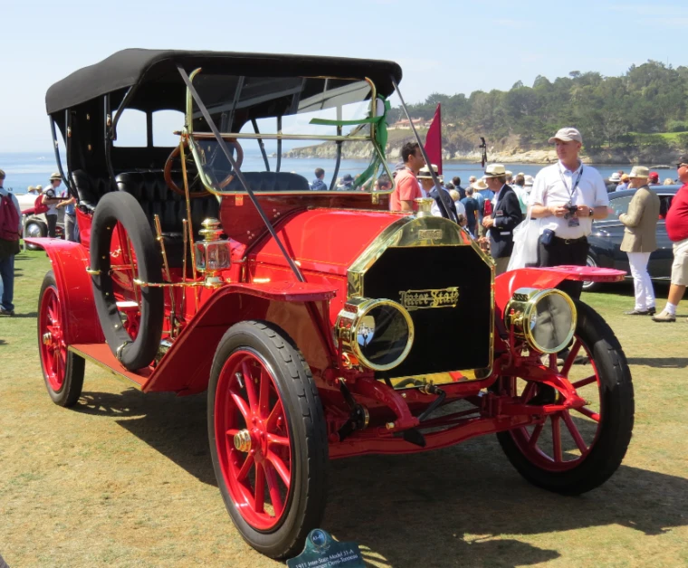 A red brass-era touring car with a black folding top stands on the lawn while spectators in summer clothes walk by, with the bay and cypress-covered point beyond (2015).