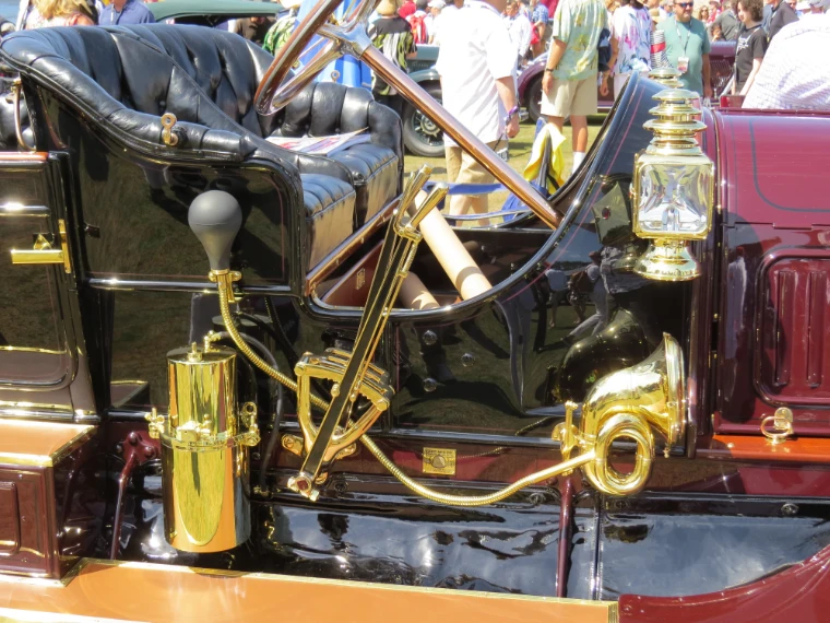 Close view of a polished brass carriage lamp, horn and gear lever on a maroon and black veteran car, with spectators blurred in the background (2015).