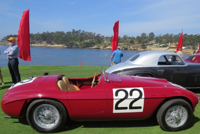 A red open-cockpit Ferrari racing car with the number 22 sits on the show lawn with the blue bay, wooded shoreline and red pennants behind it (2015).