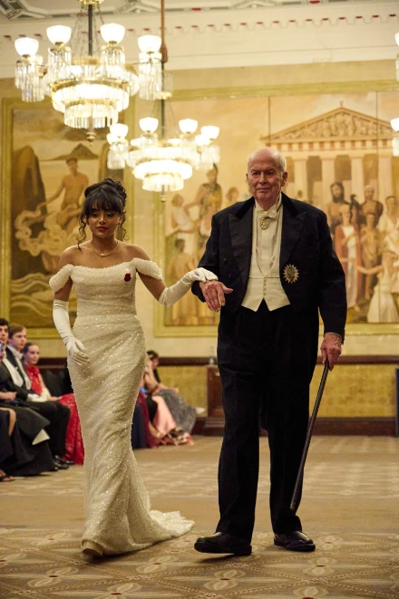 A debutante in a white gown is brought forward on the arm of a gentleman in white tie, beneath chandeliers. Queen Charlotte’s Ball, One Whitehall Place, London, 2025, in the season calendar of The Mansa Court
