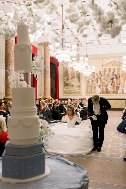 A tall white tiered cake in the foreground, a debutante’s curtsey beyond it. Queen Charlotte’s Ball, London, 2026, in the season calendar of The Mansa Court