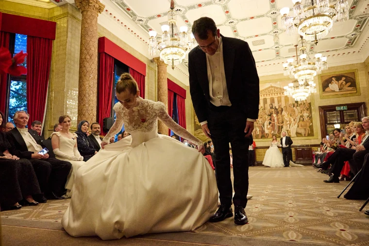 A debutante sinks into a curtsey beside her escort in white tie, beneath the murals and chandeliers of the hall, guests seated along the walls. Queen Charlotte’s Ball, One Whitehall Place, London, 2025, in the season calendar of The Mansa Court