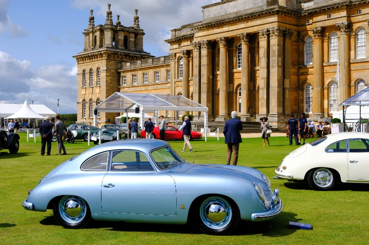 Two Porsche 356 coupés, one silver-blue and one cream, on the lawn before the columned Baroque front of Blenheim Palace, with marquees and visitors behind (2023).