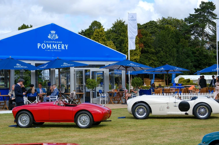 Guests seated under blue parasols outside a blue Champagne Pommery pavilion, with a red open sports car and a white racing car parked on the lawn in front (2023).