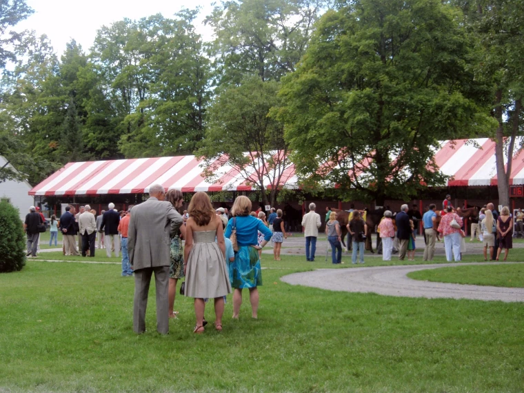 Racegoers in summer dresses and jackets gather on the paddock lawn beneath leafy trees and long red-and-white striped tents (2010).