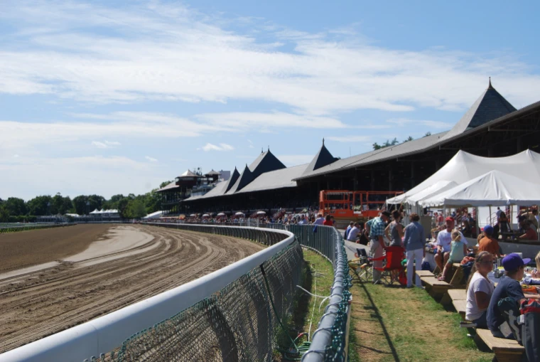 The dirt track curves past the rail toward the long Victorian grandstand with its steep slate spires, with racegoers at picnic tables and white tents along the rail under a summer sky (2009).