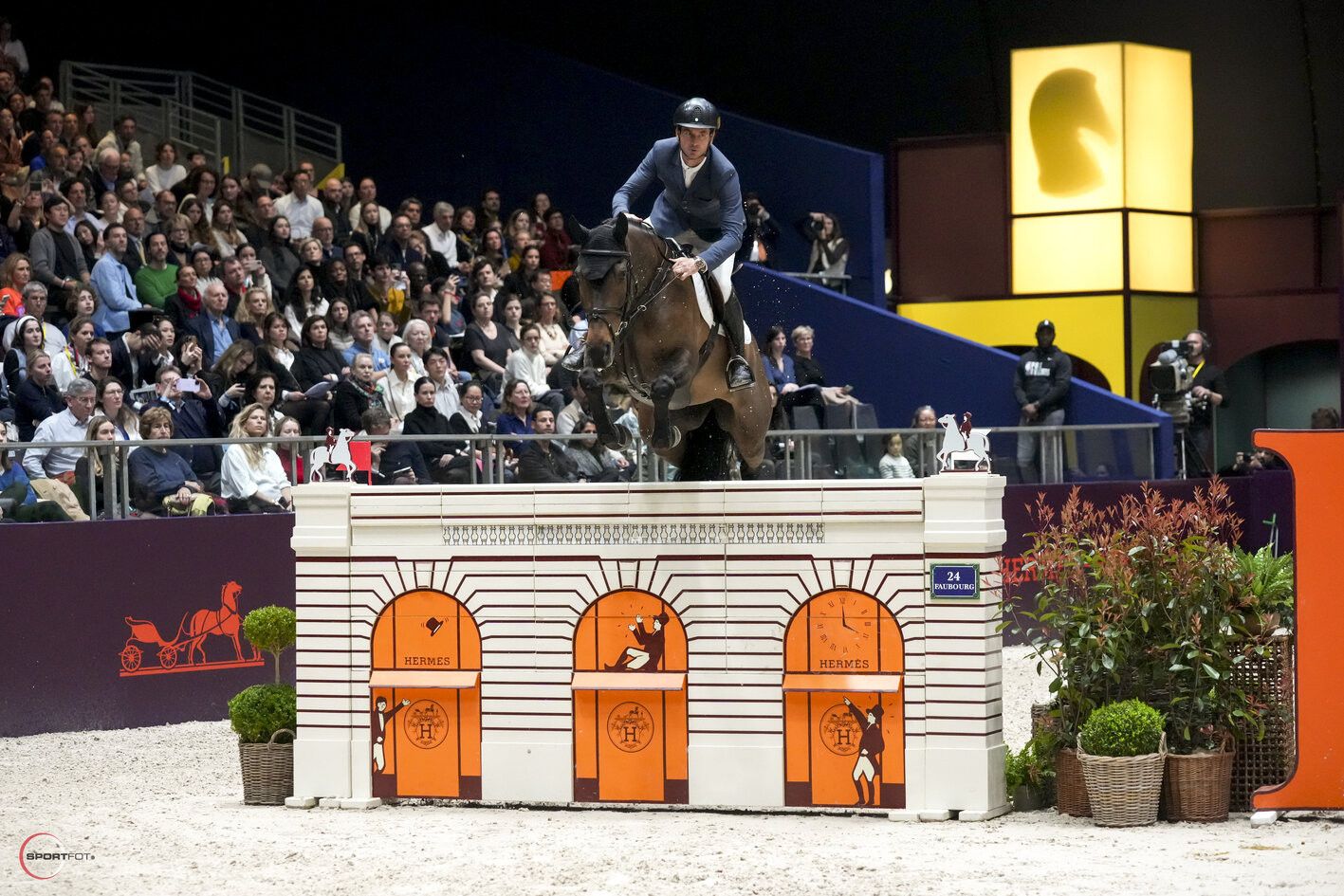 A horse and rider clearing the Hermès Faubourg shopfront fence at Saut Hermès, attended by members of The Mansa Court