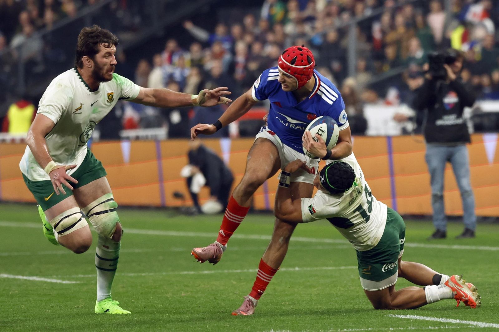 A winger in a red scrum cap carrying the ball into contact and breaking a tackle near the line under floodlight