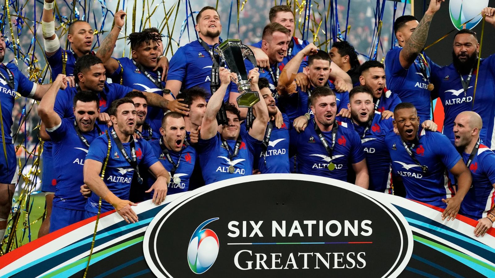 The France squad in blue lifting the Six Nations Championship trophy under gold and blue streamers on the presentation stage