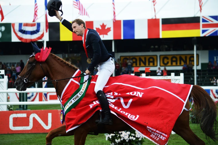 A rider in a red champion’s rug raises his helmet on a lap of the ring, with national flags behind (2013).