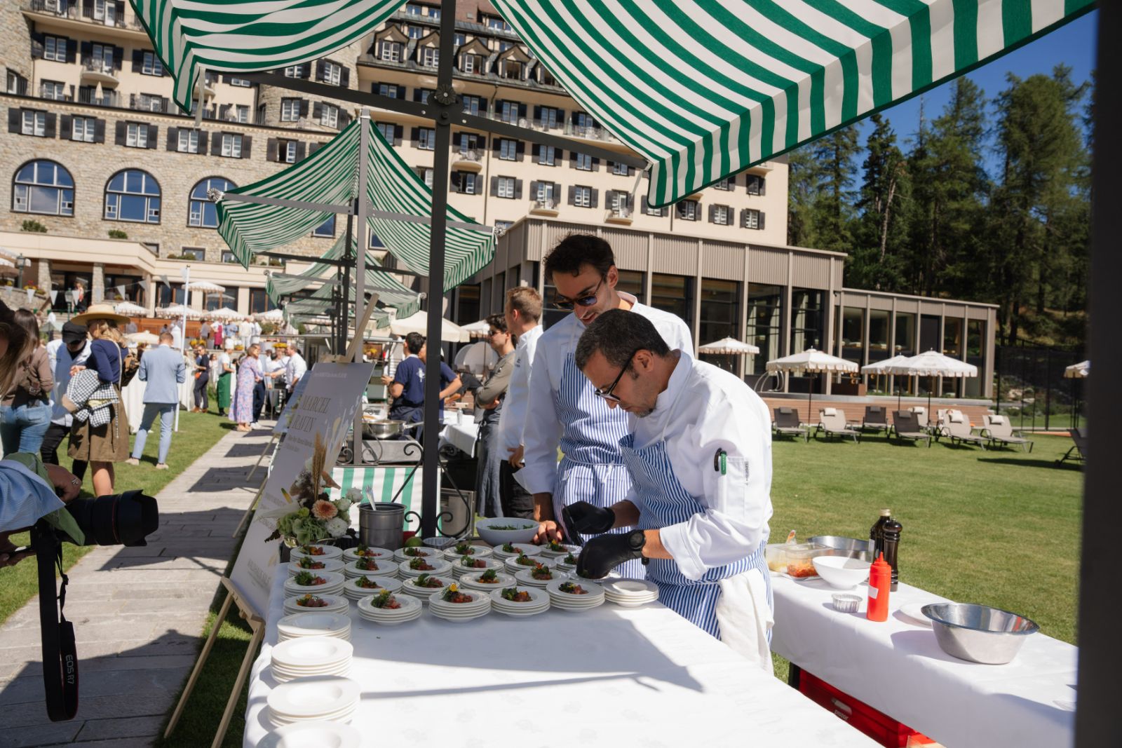 A guest chef finishing rows of tartare plates under a green-striped awning on the hotel lawn at the St. Moritz Gourmet Festival
