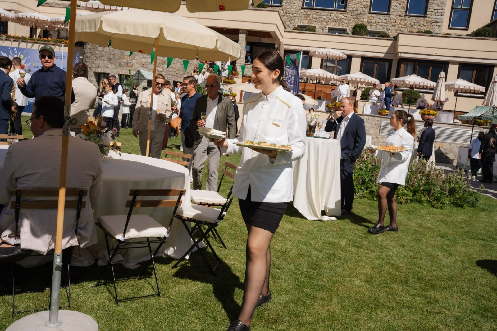 Staff in white jackets carrying trays of canapés across the hotel lawn at the St. Moritz Gourmet Festival garden party