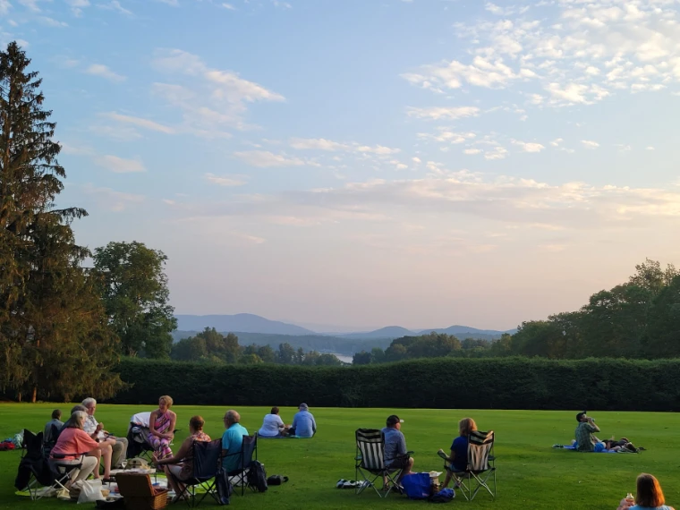 Picnickers in lawn chairs relax on a wide green lawn edged by a tall hedge, looking out over a lake and the blue Berkshire hills under a soft evening sky (2021).