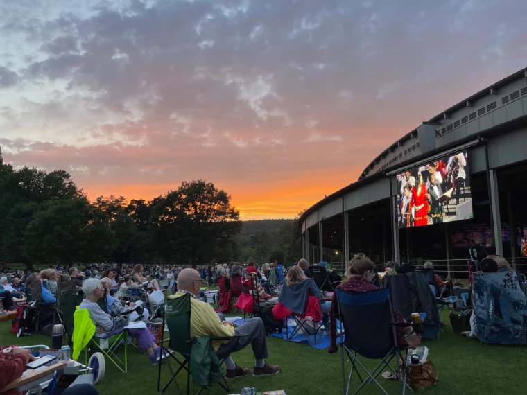 An orange sunset glows behind trees as a crowd in lawn chairs watches a concert on a large screen on the side of the Music Shed (2022).