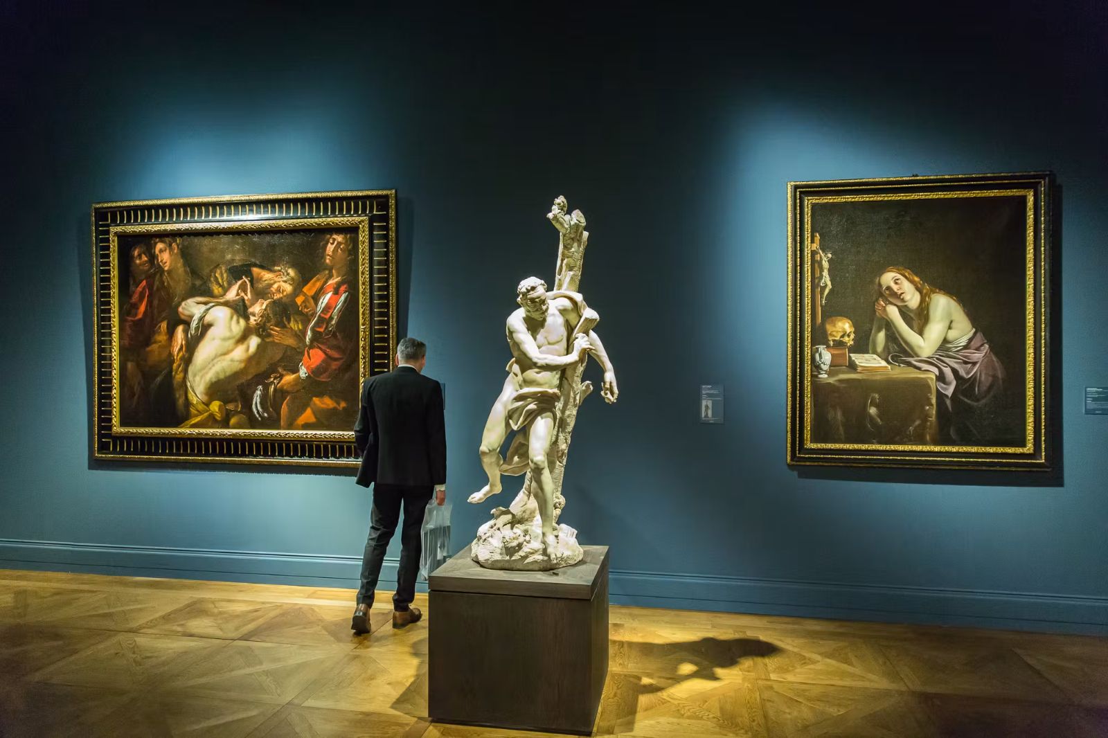 A visitor studying old master paintings and a marble sculpture on a blue-walled stand at TEFAF Maastricht, an engagement in The Mansa Court season