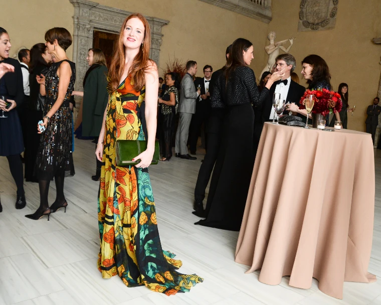 A guest in a long printed gown stands among guests in black tie and evening dress beside a draped cocktail table (2013).