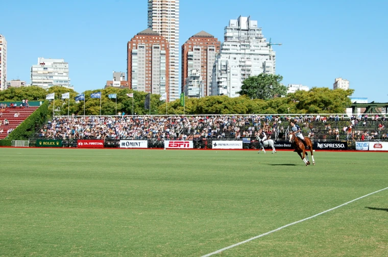 Two players gallop along the main ground in front of a packed stand lined with sponsor boards, with Palermo’s tower blocks above (2012).