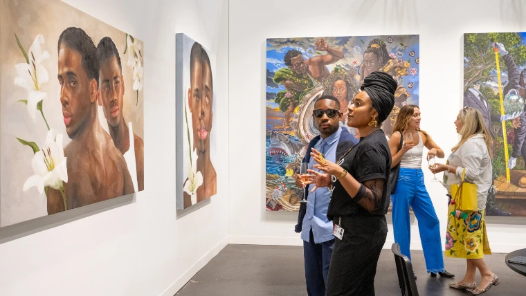 Visitors talk in a white gallery booth hung with large figurative paintings, including one of two men with white lilies (2025).