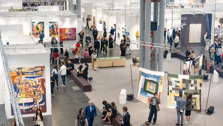 Seen from above, visitors walk and sit along a wide aisle between white gallery booths hung with paintings and quilts (2025).