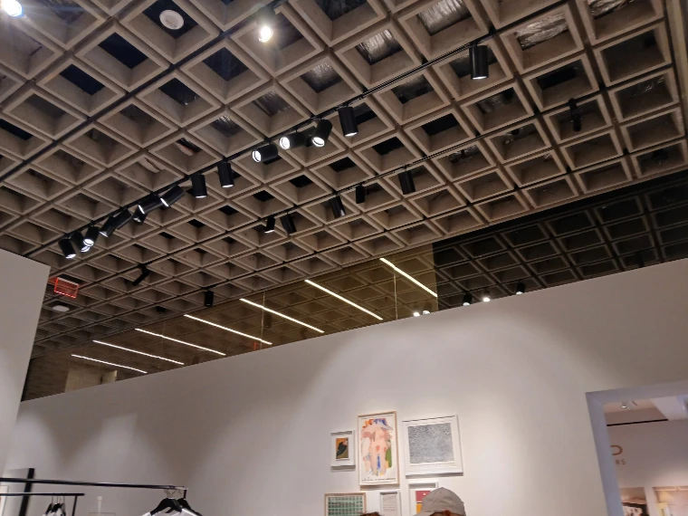 A gallery in the Breuer building under its coffered concrete ceiling and spotlights, framed works on a white wall below (2025).