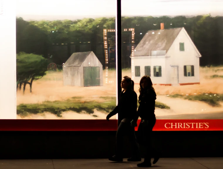 Two passers-by in silhouette walk at night past a lit Christie’s window filled with a large reproduction of a painting of white clapboard houses in dune grass, the Christie’s name in white on a red band below (2012).