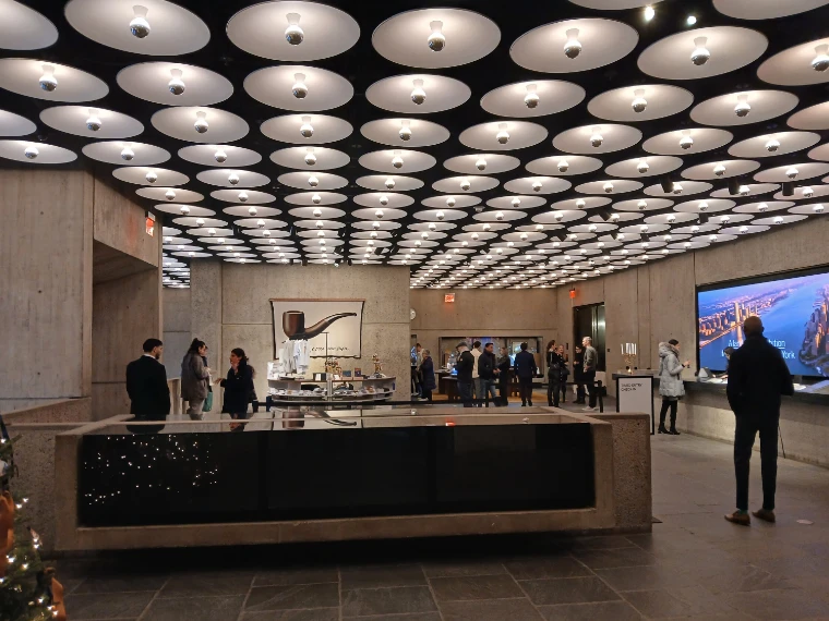 The lobby of 945 Madison Avenue under its ceiling of round lamps, visitors by the long reception desk, a pipe sculpture on a plinth and a large screen on the wall (2025).