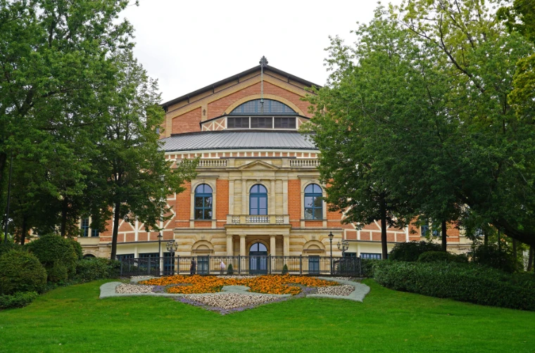 The red-brick and sandstone Festspielhaus framed by leafy trees, with a flower bed in orange and violet on the lawn before it (2020).