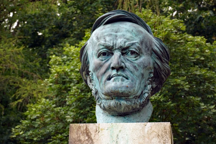 A weathered bronze bust of Richard Wagner on a stone plinth against dense summer foliage (2020).