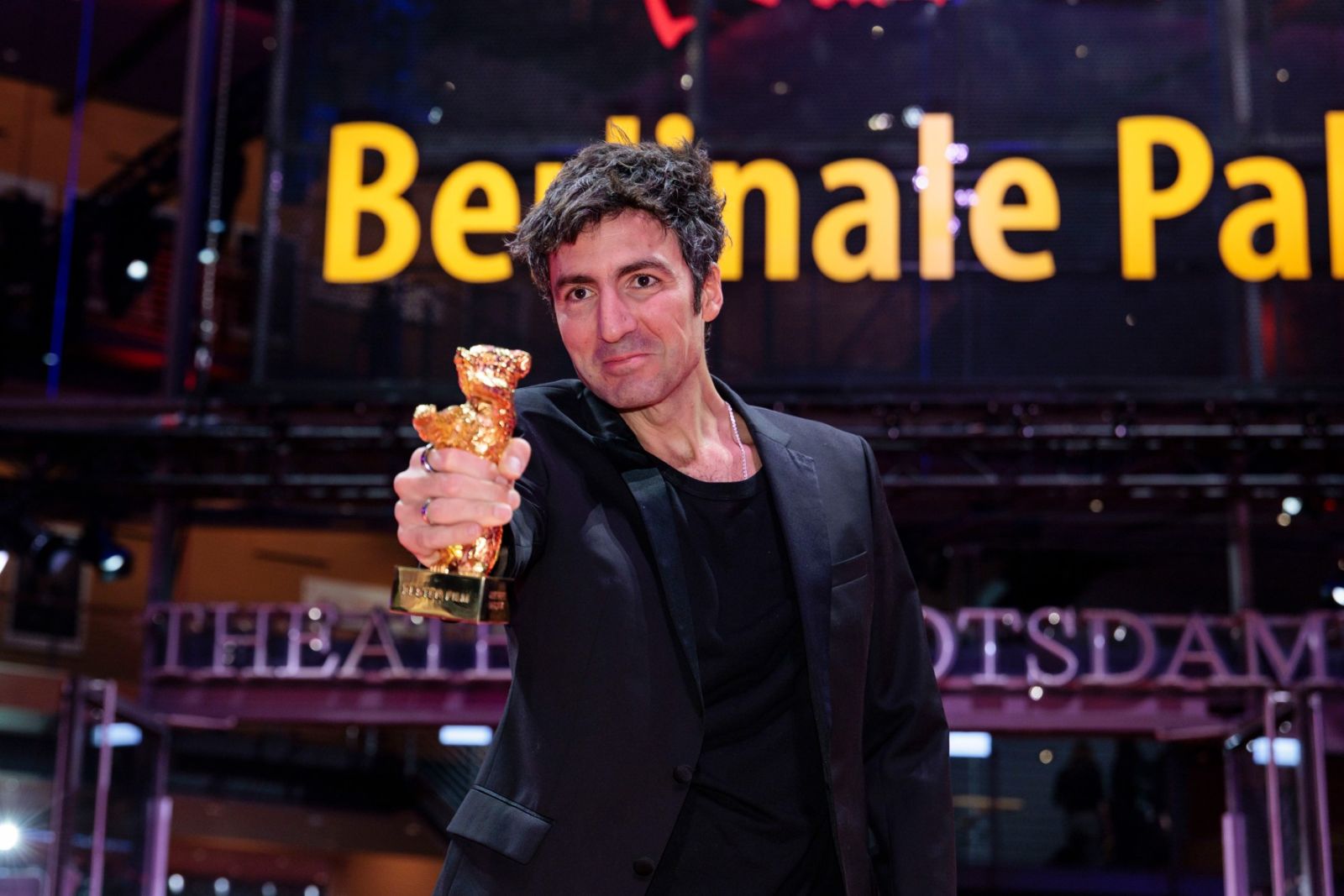 A winning director holding the Golden Bear out to the cameras outside the lit Berlinale Palast at The Berlinale