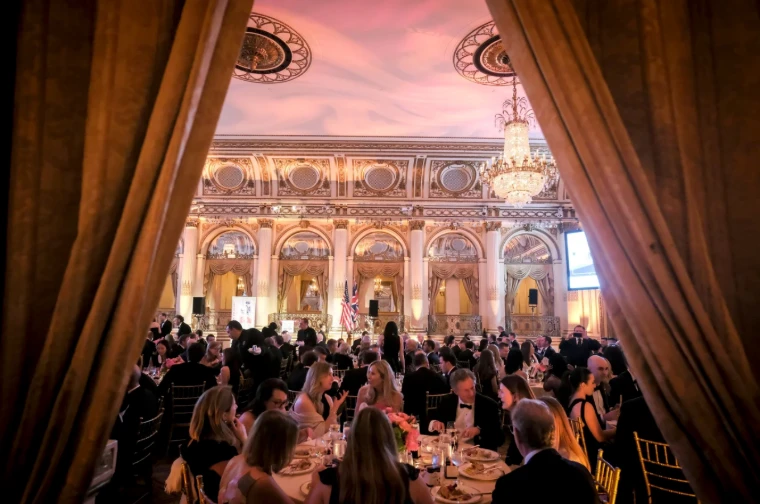 The gilded ballroom at dinner seen between parted curtains, the Union and American flags on the stage, at The British Ball in New York, an April engagement in the season calendar of The Mansa Court