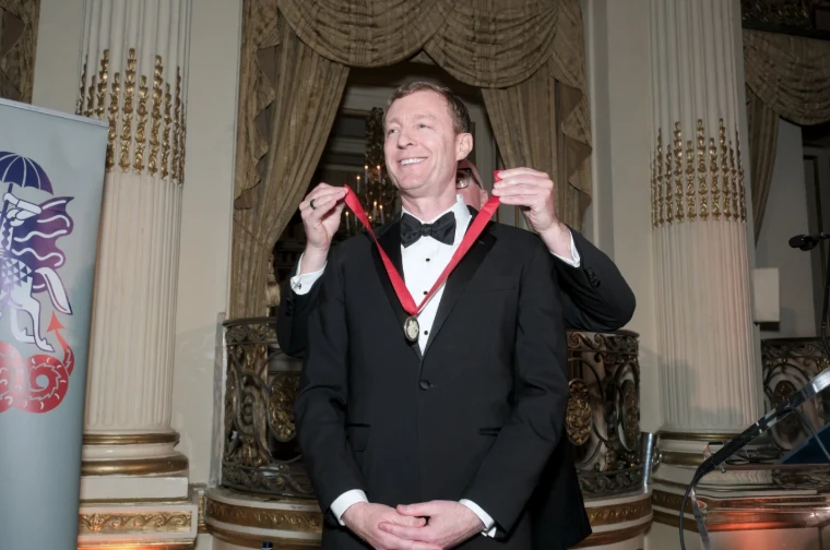 An honoree in black tie receiving a medal on a red ribbon before the gilded columns of the ballroom at The British Ball in New York, an April engagement in the season calendar of The Mansa Court