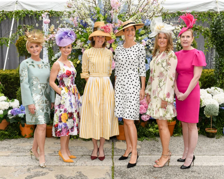 Six guests in stripes, polka dots and florals, each in a hat or fascinator, before a wall of cherry blossom and hydrangea. The Central Park Conservancy Hat Luncheon in the Conservatory Garden, New York, in the season calendar of The Mansa Court