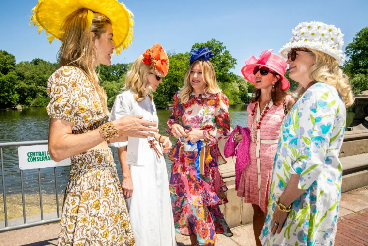 Five guests in bright prints and hats, from yellow feathers to white daisies, talk beside the lake in Central Park. The Central Park Conservancy Hat Luncheon in the Conservatory Garden, New York, in the season calendar of The Mansa Court