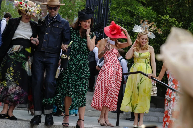 Guests hold their hats against the wind as they come down the steps from the garden gates, arm in arm. The Central Park Conservancy Hat Luncheon in the Conservatory Garden, New York, in the season calendar of The Mansa Court