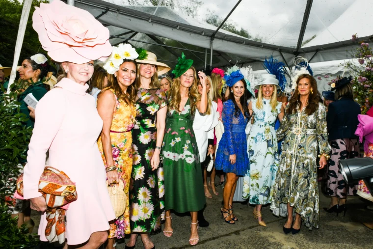 Guests in floral dresses and sculptural hats, one a vast pink paper rose, gather beneath the clear tent on a rainy spring day. The Central Park Conservancy Hat Luncheon in the Conservatory Garden, New York, in the season calendar of The Mansa Court