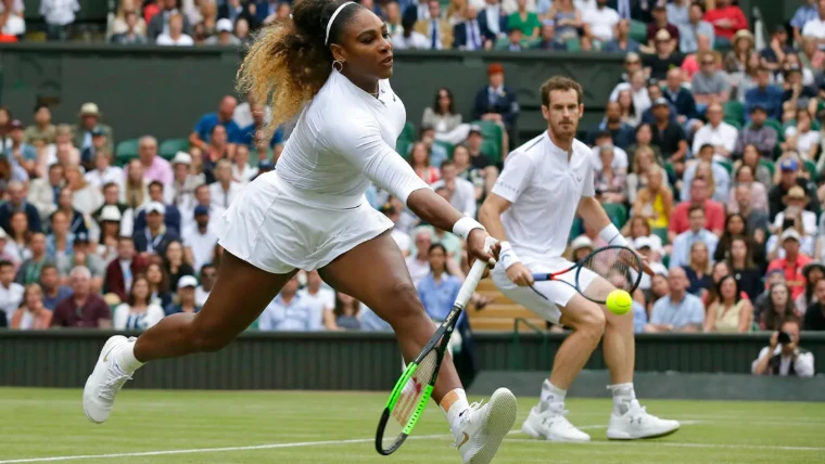 A mixed doubles pair in tennis whites stretching for a volley on the grass before a full stand at The Championships, Wimbledon, London, an early-July engagement in the season calendar of The Mansa Court