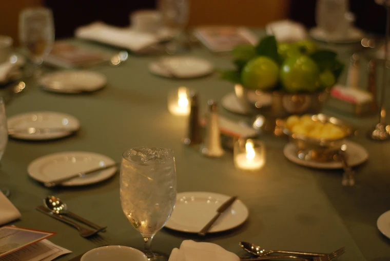 A gala dinner table laid with plates, silver, iced water glasses and small candles, a bowl of green apples at its centre, softly out of focus (2009).