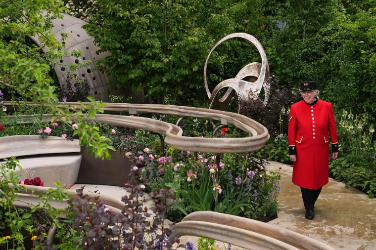 A Chelsea Pensioner in scarlet walks through a show garden of irises, pinks and bronze sculpture. The RHS Chelsea Flower Show in the grounds of the Royal Hospital Chelsea, London, in the season calendar of The Mansa Court