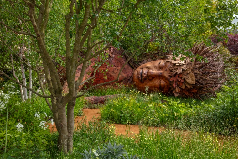 A great carved wooden figure lies asleep beneath the trees of a woodland show garden. The RHS Chelsea Flower Show in the grounds of the Royal Hospital Chelsea, London, in the season calendar of The Mansa Court