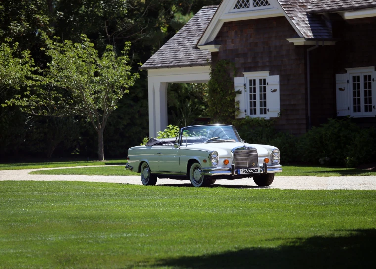 A pale classic Mercedes-Benz cabriolet with its top down stands on a gravel drive before a cedar-shingled house and a mown lawn (2015).